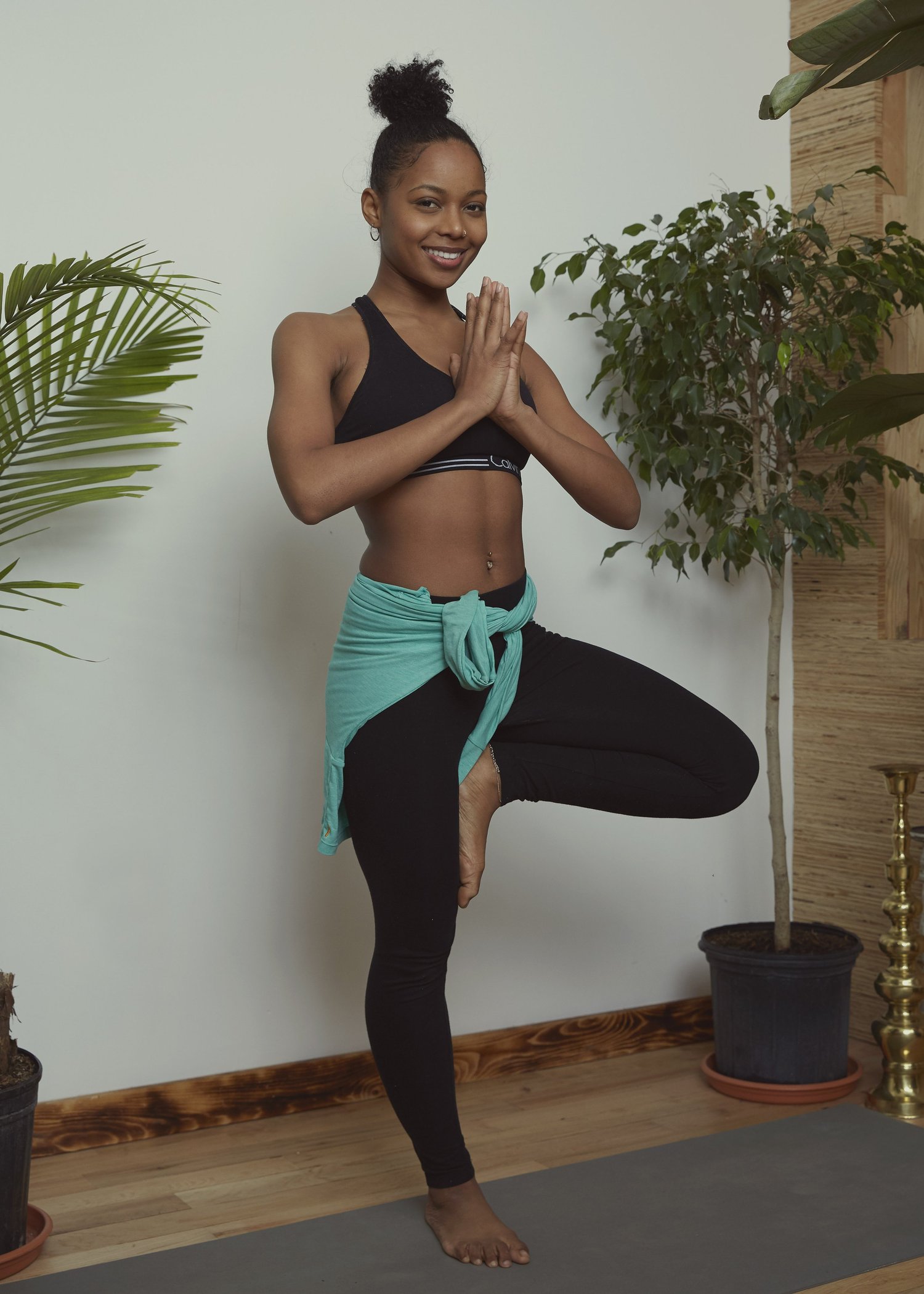 A student in tree pose against the studio's white wall, framed by the studio's potted plants