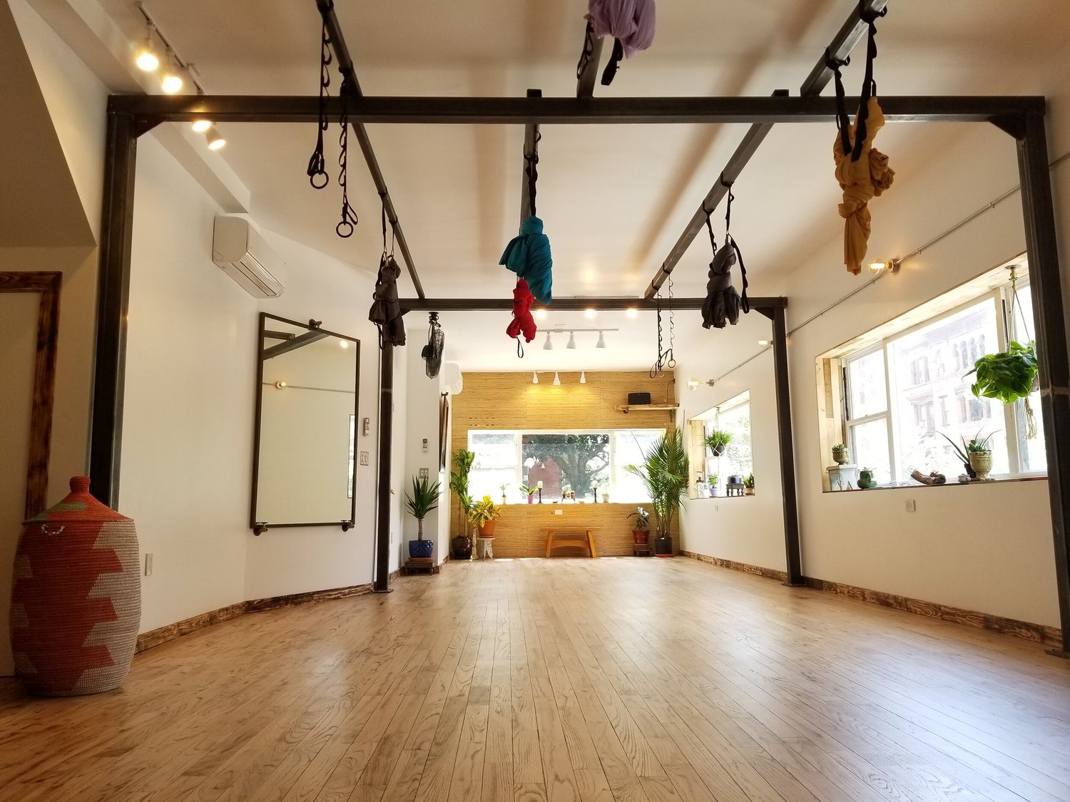 Interior of And Yoga Studios' Nostrand Ave room — pale wood floor, white walls, plants, and the steel rig with aerial hammocks knotted overhead